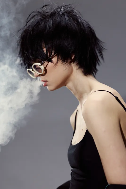 Young woman with short black hair wearing glasses and black tank top exhaling vapor in a moody studio setting