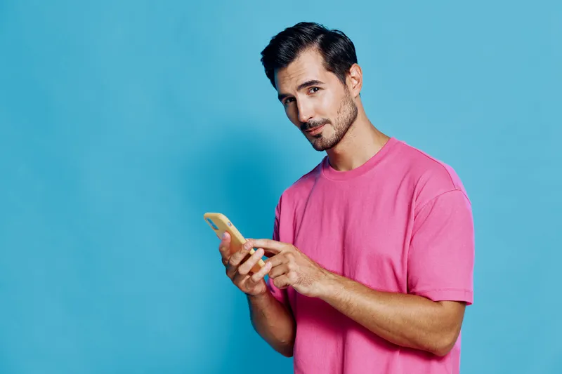 Smiling man in a casual pink t-shirt holding a smartphone, looking confidently at the camera against a vibrant blue background. Lifestyle and technology concept.