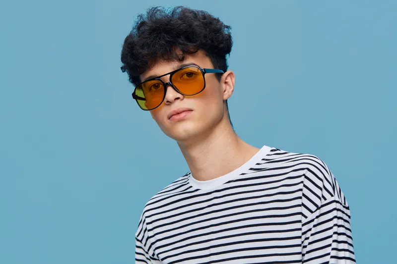 young man wearing yellow sunglasses and striped shirt stands against blue background with calm expression and short curly hair, casual style portrait