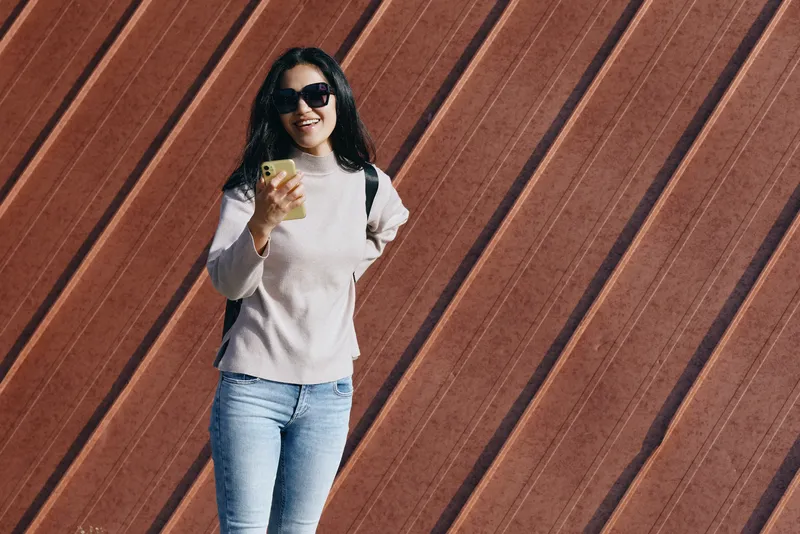 Female in casual attire stands outdoors against a wooden wall, smiling with sunglasses, holding a smartphone, capturing everyday moments, style, travel, and social sharing in a bright urban setting.