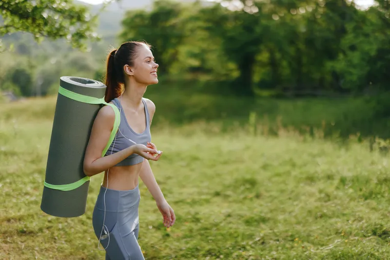 Young woman carrying yoga mat in a serene green landscape, embodying wellness, fitness, and outdoor activity