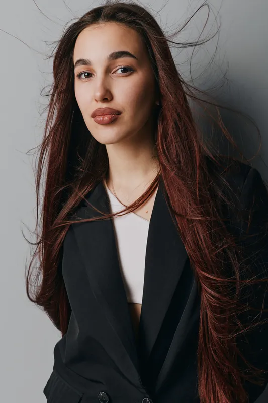 Young woman with long brown hair wearing black blazer and white top standing against gray background with hair flowing, confident expression and natural makeup in studio portrait.