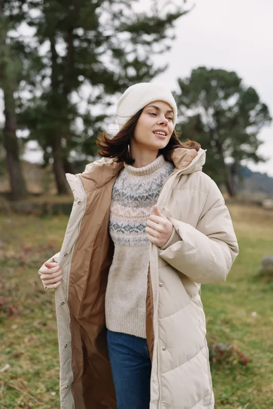 Woman in a padded coat enjoying a windy outdoor day, wearing a knit sweater and beanie in a park with trees