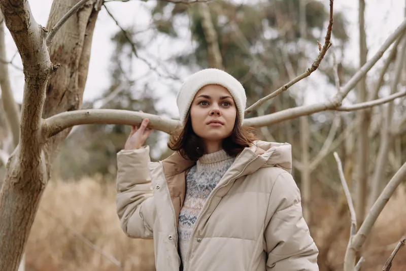 Woman in a beige winter coat stands outdoors among bare trees, capturing a calm, serene moment in natural light with a cozy, stylish vibe and reflective mood.