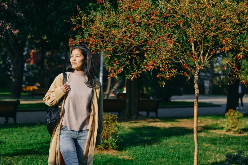 Woman walking in a sunlit park during autumn, wearing a long beige coat and sweater, carrying a bag, amid red and orange foliage, green grass, and quiet paths, conveying relaxed outdoor lifestyle and