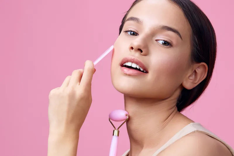 Young woman applying skincare with facial roller on pink background, promoting beauty, confidence, and self care