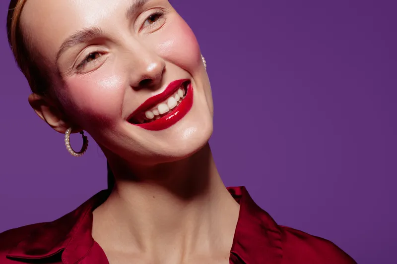 Smiling woman with bright red lipstick and hoop earrings poses against purple background. Close-up portrait with smooth skin and shiny makeup. Concept of beauty, fashion, and confidence.