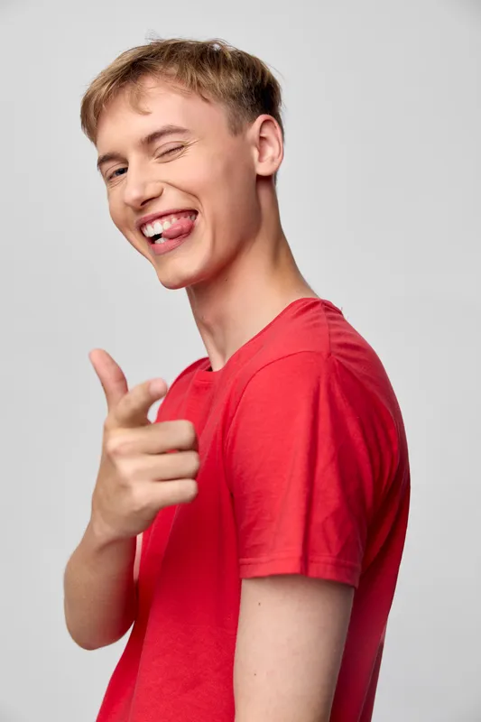 Smiling man in red shirt pointing playfully at the camera, joyful expression, casual studio setup, friendly engagement, lively pose, bright background