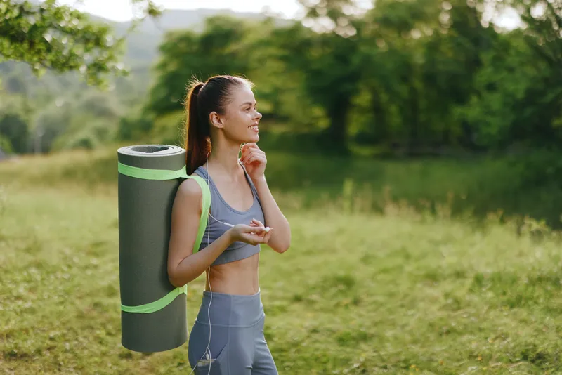 Young fitness woman enjoying nature with yoga mat, wearing sporty outfit, smiling in green outdoor setting, healthy lifestyle concept