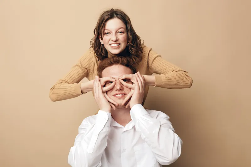 Happy young woman in brown sweater playfully covers eyes of smiling man in white shirt. Studio portrait on beige background. Concept of friendship, fun, joy, and positive emotions.