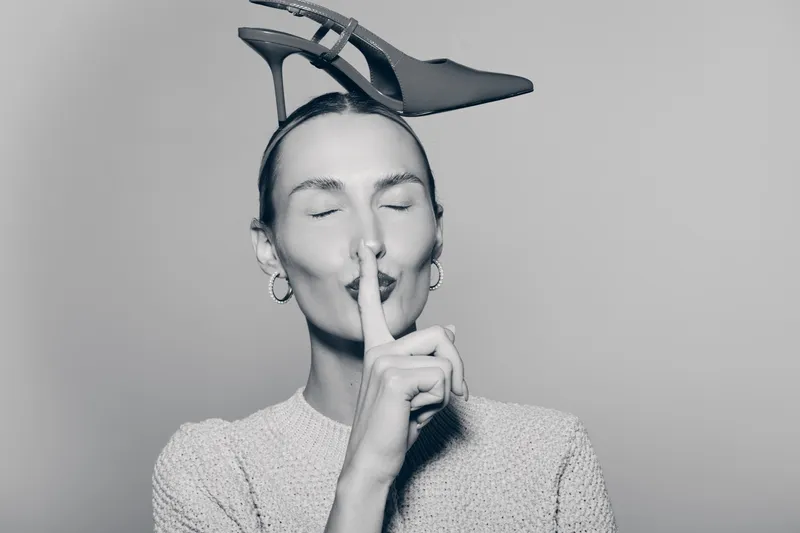 Light skinned Caucasian woman with short hair holds finger to lips in a shushing gesture while balancing a high heel shoe on her head in studio.