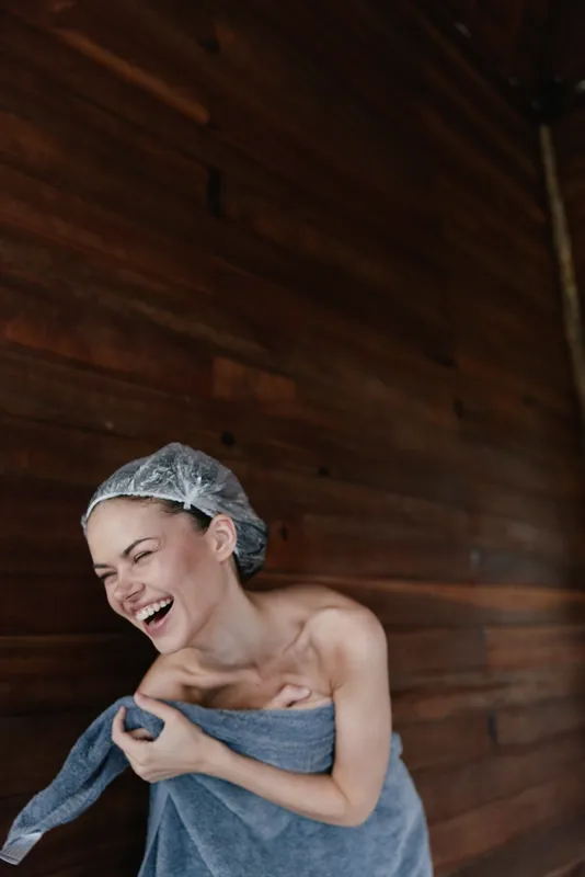 laughing woman wrapped in towel and wearing hair cap stands against wooden wall, expressing joy and relaxation in warm spa atmosphere, natural light enhances her smile.