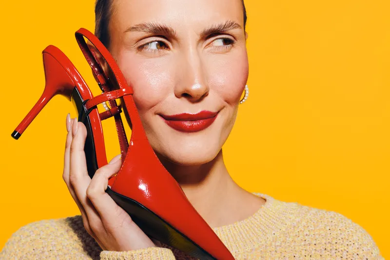 Close-up of a smiling woman holding red high heel shoes near her face against a bright yellow background. Concept of fashion, style, beauty, and confident female model with vibrant colors.