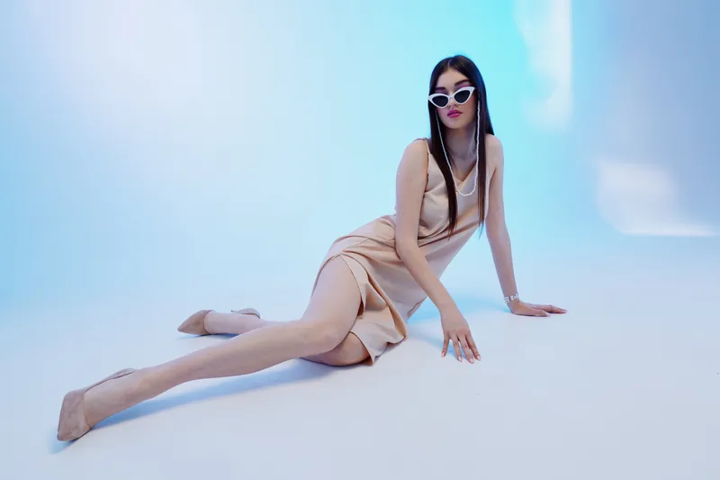 Elegant woman beauty fashion posing on floor wearing beige dress and white sunglasses in studio with soft blue lighting. Concept for style, glamour, and modern elegance.