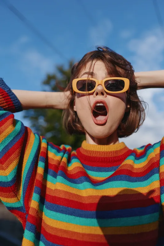 Woman in yellow sunglasses and rainbow sweater surprised with open mouth in a colorful portrait outdoors. Short hair, expressive fashion vibe and playful mood against blue sky.