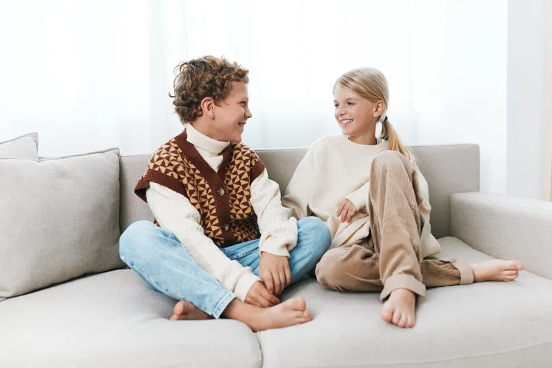 Two children sit on a light sofa, facing each other with warm smiles, cozy sweaters, and relaxed posture, sharing a friendly moment in a bright living room.