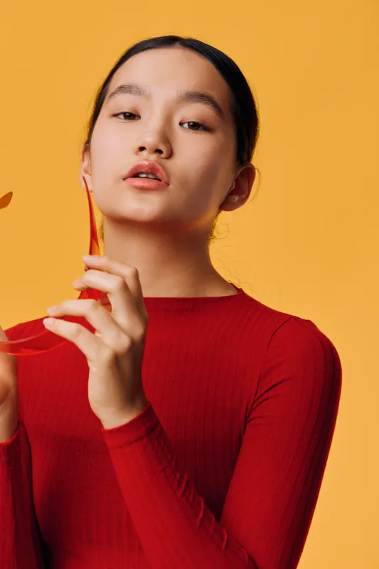 Young woman in red sweater holding dried leaves with delicate hands on orange background. Portrait with natural makeup and calm expression in minimalistic style.