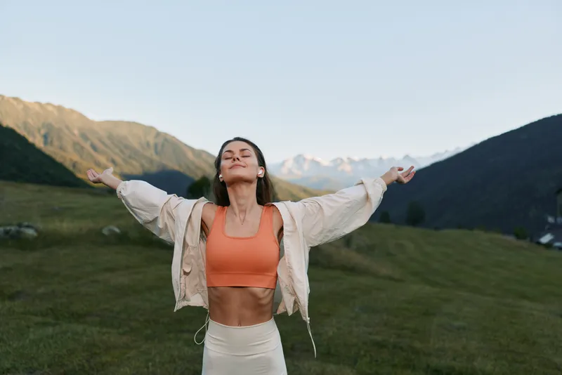 A cheerful woman stands in a grassy valley with distant mountains. She wears athletic attire, arms raised, eyes closed, and a content smile, expressing freedom, vitality, and a sense of wellbeing in