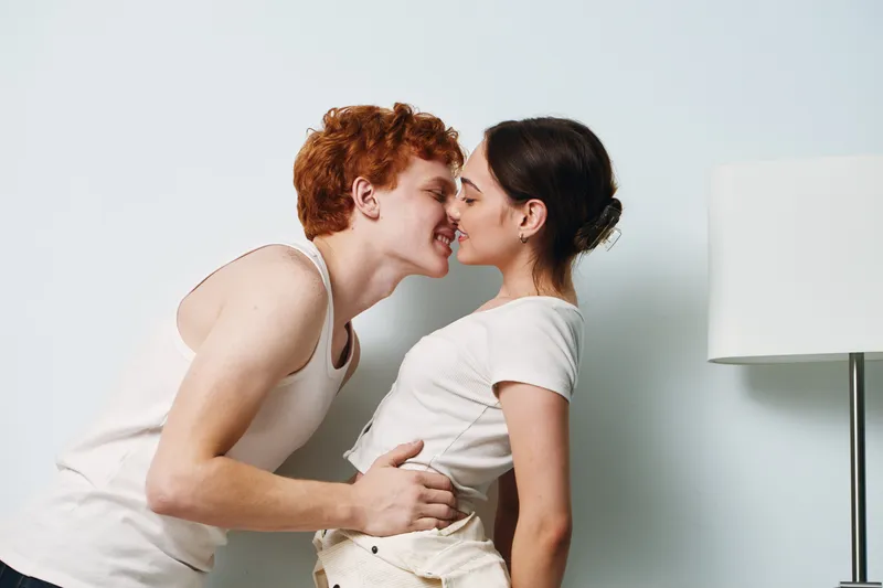 Couple in casual clothes sharing intimate moment touching noses and smiling indoors near white wall and lamp, affection and love between young people at home.