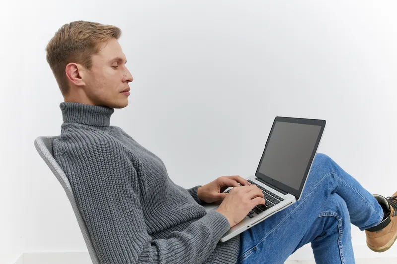 Man sitting on chair using laptop computer with gray turtleneck sweater and jeans, working or studying in modern minimalist office environment, concept of remote work and digital technology, indoor sh