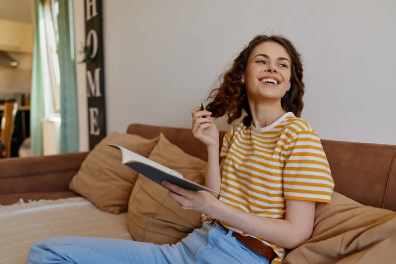 A smiling woman sits on a couch with a notebook in a bright home, conveying casual lifestyle and cozy atmosphere in a comfortable living space.