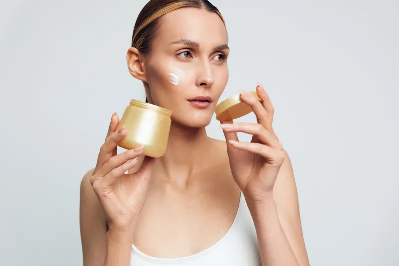 Smiling woman applying white face cream on her cheek. Close-up on a light background. Concept of skincare, wellness, and self care. Soft studio lighting creates a calm, radiant mood.
