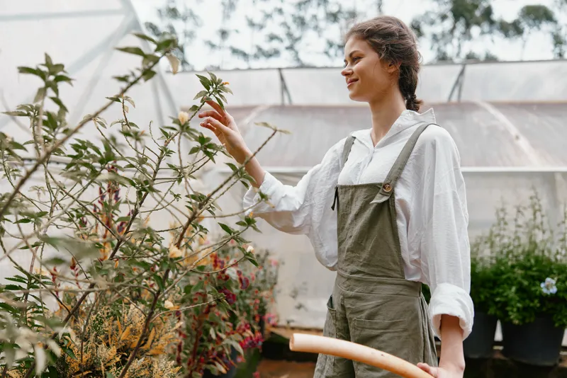 Young woman in overalls tending to plants in a greenhouse, expressing joy and connection to nature, ideal for concepts of gardening, sustainability, and nature