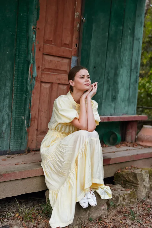 Woman thoughtful sitting on a wooden porch in a flowing yellow dress, gazing into the distance, capturing a calm outdoor moment with soft light and relaxed mood.