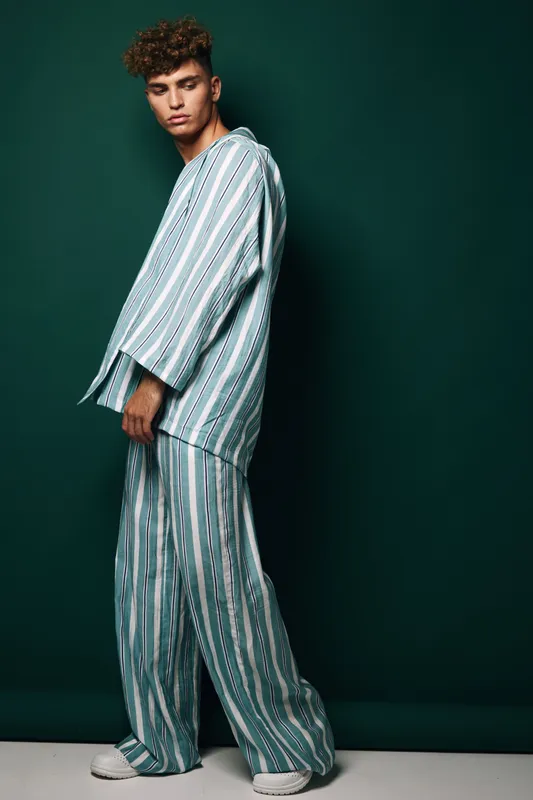 Young man wearing striped loose pajamas standing against dark background with casual posture and white sneakers for comfortable homewear style.