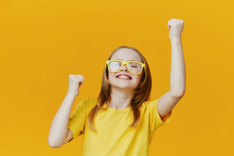 Happy girl celebrating against a vibrant yellow background, wearing yellow glasses and a t shirt, expressing joy and excitement in a cheerful mood
