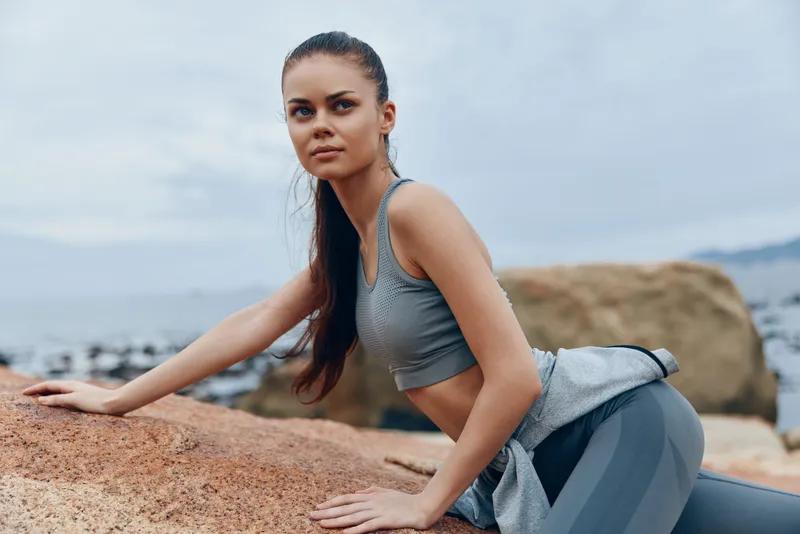 Athletic woman in gray sportswear practicing yoga on rocky beach, expressing determination, with a serene ocean background and cloudy sky