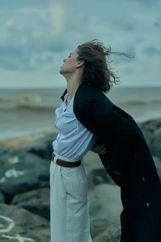 Windblown woman in smart attire on a windy coast, stands with hands on hips, rocks and ocean behind, conveying confidence and motion.