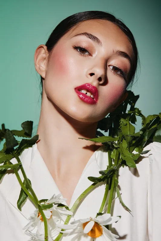 Young woman with glowing radiant skin and glossy red lips poses gracefully with white daisies on a light green backdrop. Concept of beauty creativity and fashion glamour.