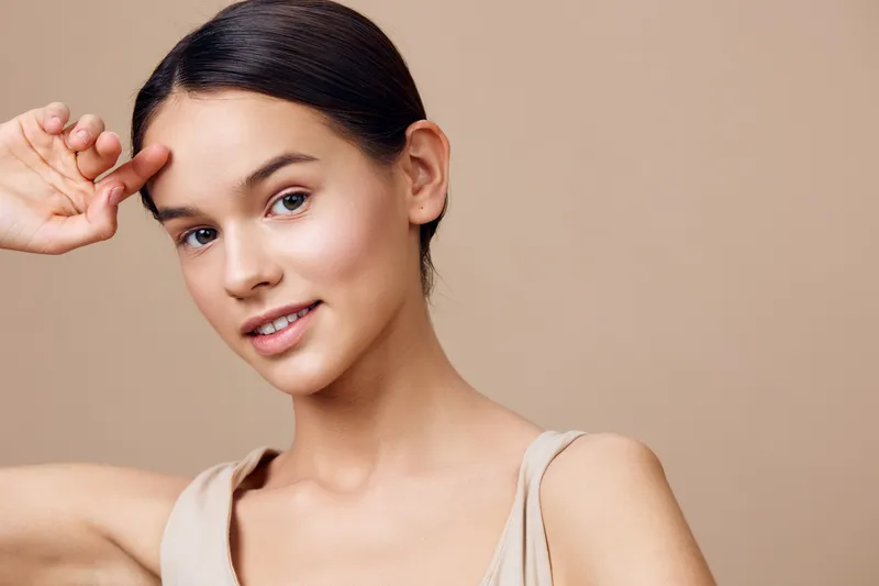 Young woman with medium skin tone smiling softly, wearing a neutral colored top, embodying beauty and confidence against a beige background