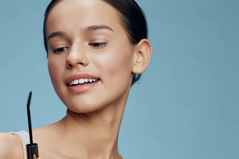 Young woman applying mascara with a gentle smile, showcasing beauty and confidence against a soft blue background Perfect for beauty, makeup, and skincare themes