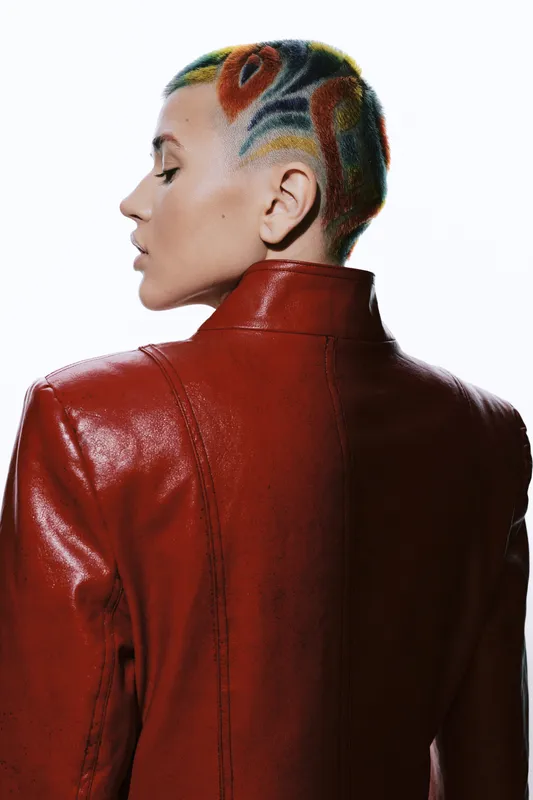 Woman with colorful shaved hair wearing a bright red leather jacket, showcasing bold style and modern fashion with artistic hair design in a studio setting.