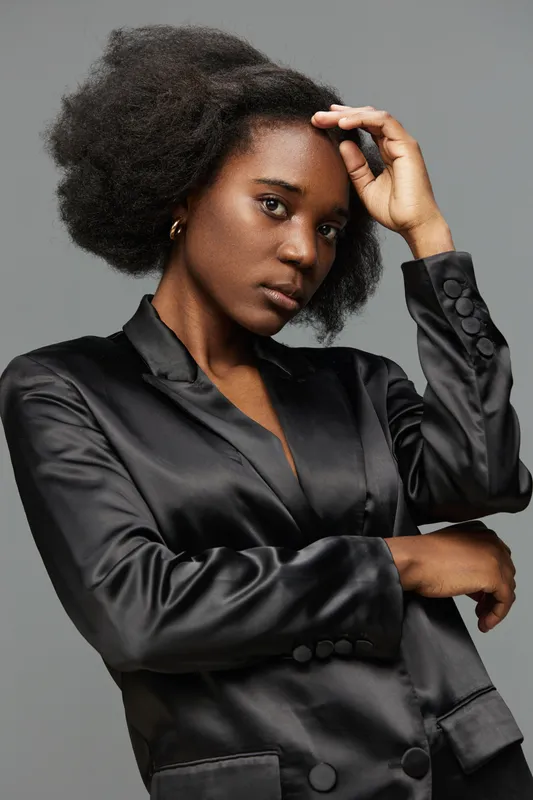 Elegant African woman in a black satin blazer poses confidently against a minimalist gray background, showcasing beauty and self expression