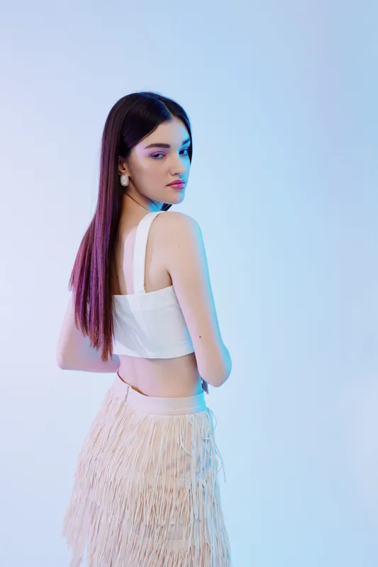 Young woman with long dark hair wearing a white crop top and fringed skirt poses with a serious expression on a light blue background. Concept of beauty fashion style.