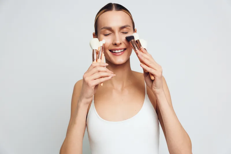 Young woman with clear skin holding makeup brushes near her face with closed eyes on white background. Concept of beauty, skincare, cosmetics, and natural care routine.
