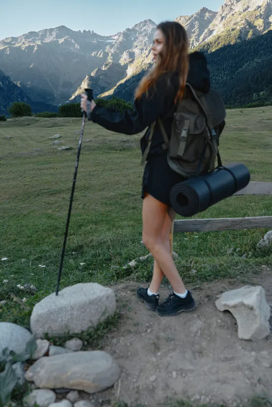 A female hiker with backpack and trekking poles stands on a rocky trail amid alpine mountains, enjoying an outdoor adventure under clear skies, conveying energy and exploration.