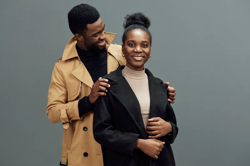 Happy couple posing together in stylish winter outfits the woman wears a black coat and the man a beige trench coat against a grey background