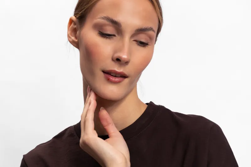 Portrait of a young woman with natural makeup wearing a dark top, looking down with a soft expression. Concept of beauty, calm, and minimalism in studio lighting.