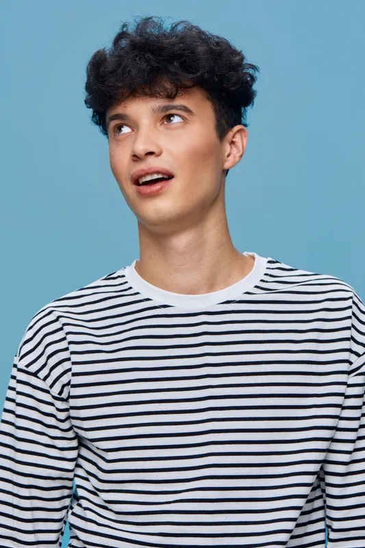 young man wearing striped shirt looking up with curious expression against plain blue background, casual style, curly hair, indoor portrait, modern fashion