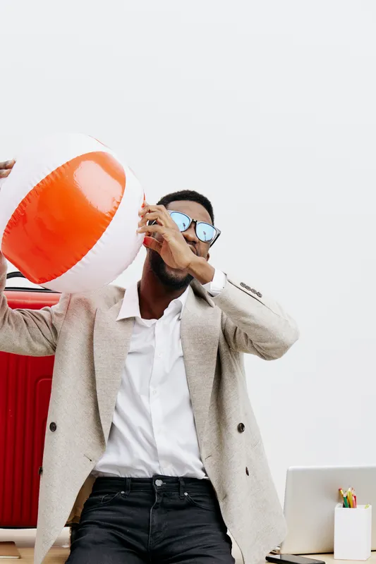 Happy young man with sunglasses and stylish beige blazer holding a colorful beach ball in a modern office environment with minimal decor, white background, and creative workspace accessories. People l
