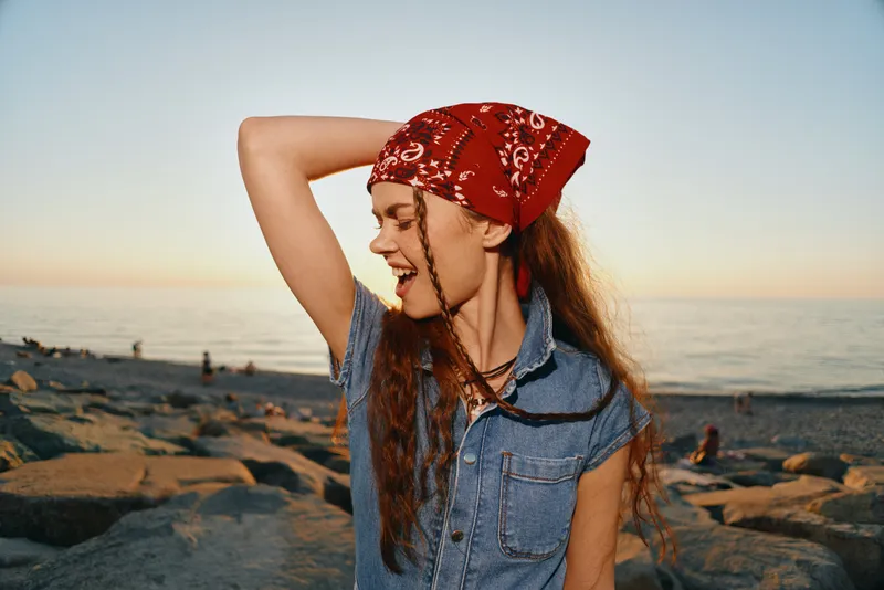 Lifestyle woman in modern boho-western denim street style with a red bandana enjoys sunset by the sea, captured in warm film color and relaxed natural mood.
