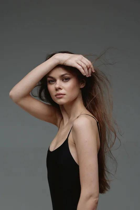 Young woman with long hair posing in black dress against gray background, natural beauty, confident, soft lighting.