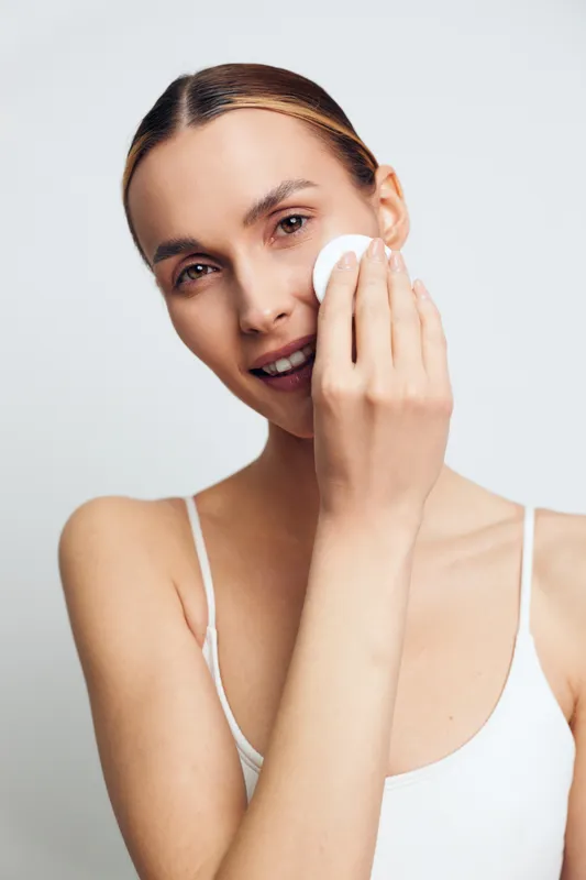 Young woman with clear skin gently cleansing her face using a cotton pad, soft natural light, minimal makeup, white background, skincare routine concept