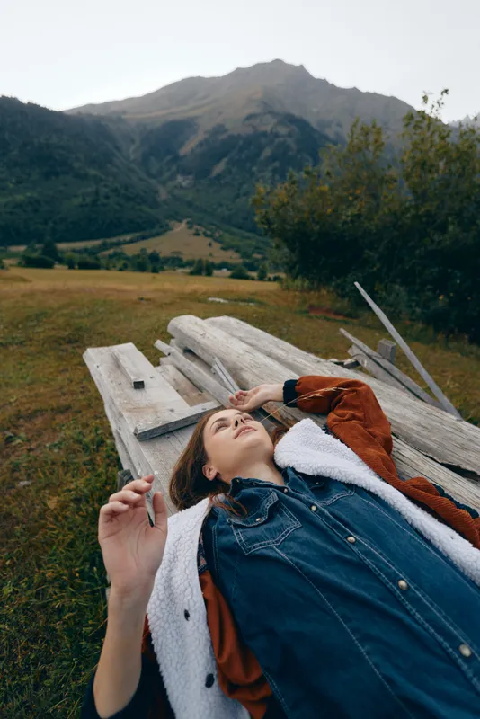 Woman lying on a wooden bench in a meadow near mountains, portrait capturing a relax outdoors moment in nature, casual denim jacket and headphones for a peaceful contemplative scene
