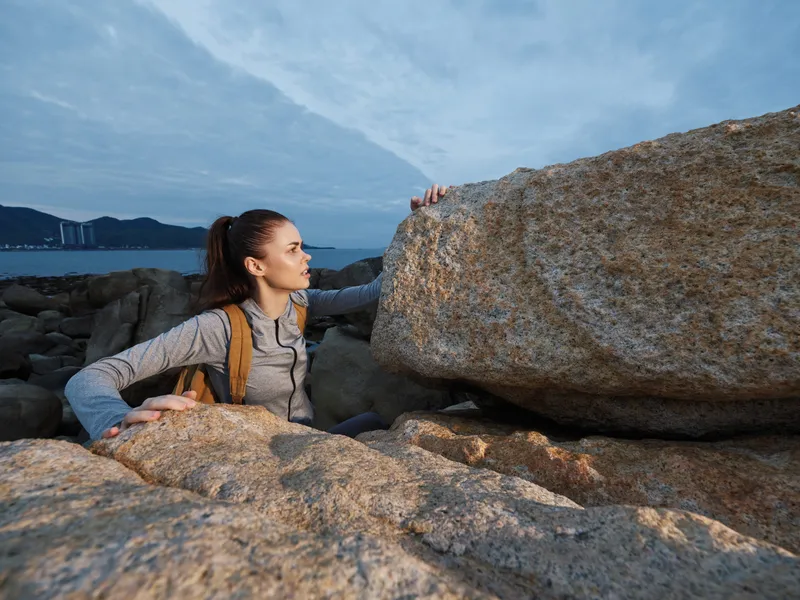 Young female hiker exploring rocky coastline, wearing outdoor sportswear, showcasing determination, against a serene seaside background with cloudy sky