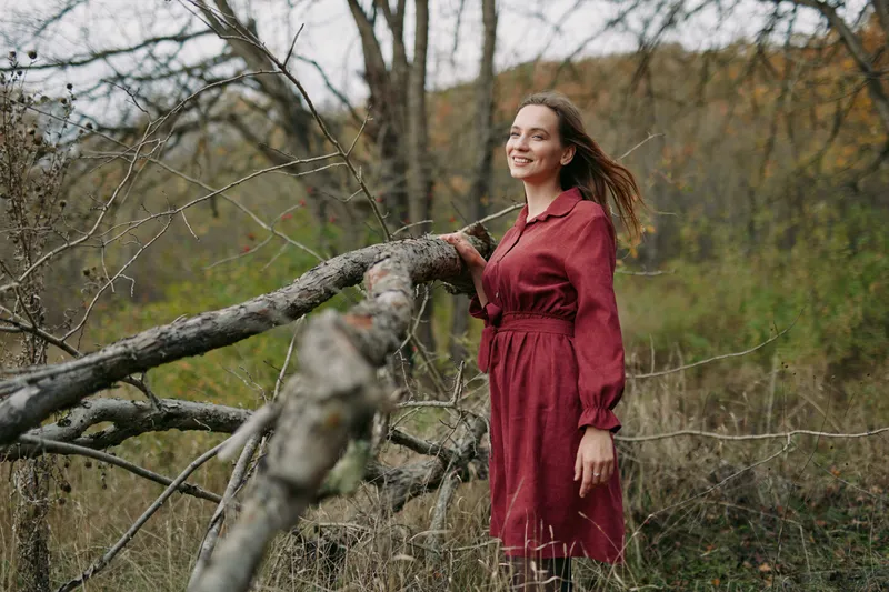 A portrait of a smiling woman in a red dress standing by a fallen tree in a meadow, captured with natural light to emphasize authenticity and veracity in her relaxed, genuine expression.